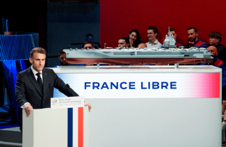 Emmanuel Macron przemawia zza mównicy z biało-czerwono-niebieskim pasem, w tle model lotniskowca oraz napis „France Libre” na ekranie.