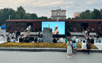 Dwa zardzewiałe wagony towarowe na placu w Kiszyniowie, przed nimi duży ekran z projekcją filmu i kamienna bryła. Widzowie siedzą na ławkach i murku przy rabacie z żółtymi kwiatami; w tle łuk z zegarem.