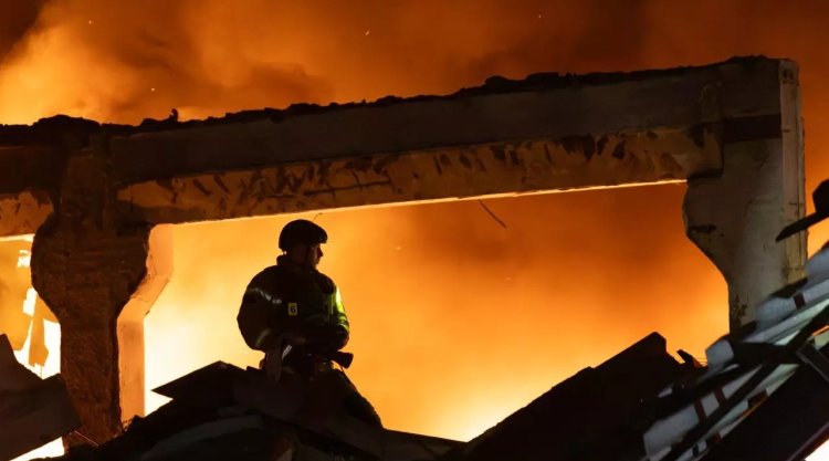A firefighter in a helmet stands against a backdrop of a burning, partially collapsed warehouse building at night; tall flames and thick smoke fill the background; the DSNS Kyiv logo appears in the upper left corner.