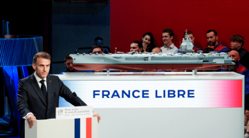Emmanuel Macron przemawia zza mównicy z biało-czerwono-niebieskim pasem, w tle model lotniskowca oraz napis „France Libre” na ekranie.